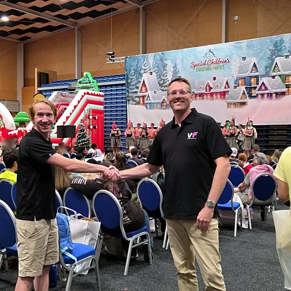 Oli Corby of VF Digital shaking hands at the Special Children's Christmas Parties, with kapa haka performers on stage behind